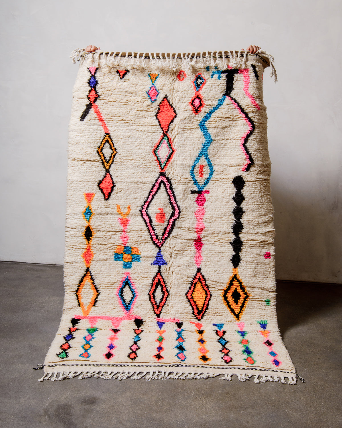 Berberlin Azilal rug made of fluffy cream wool and colourful design, held up against a white wall and cement floor.